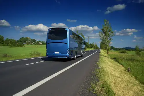Blauer Bus fährt auf Landstraße in einer ländlichen Landschaft. Dorf und Berg mit Burgruine im Hintergrund. Anreise mit dem Bus.